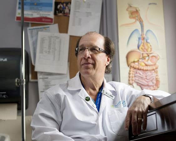 A man in white lab coat sitting next to a wall.