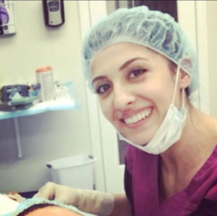 A woman in purple shirt and surgical cap smiling.